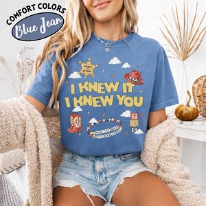 May include: A blue short-sleeve t-shirt featuring a graphic print with the text "I KNEW IT I KNEW YOU" surrounded by illustrations of a sheriff badge, a cowboy hat, boots, a beaded bracelet with "YOU'VE GOT A FRIEND IN ME" text, and blocks.
