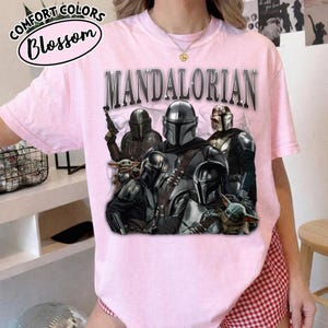 Puede incluir: Una camiseta rosa claro con un gr&aacute;fico de personajes de The Mandalorian. La palabra "MANDALORIAN" est&aacute; impresa en la parte superior en una fuente plateada met&aacute;lica. La camiseta tambi&eacute;n tiene las palabras "Comfort Colors Blossom" en un dise&ntilde;o circular.