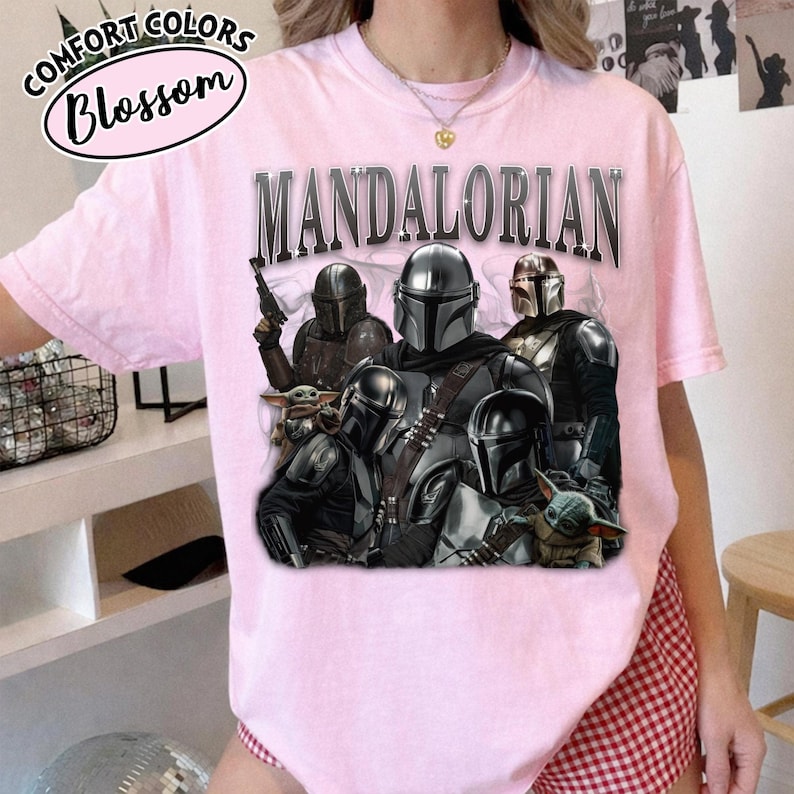 Puede incluir: Una camiseta rosa claro con un gr&aacute;fico de personajes de The Mandalorian. La palabra "MANDALORIAN" est&aacute; impresa en la parte superior en una fuente plateada met&aacute;lica. La camiseta tambi&eacute;n tiene las palabras "Comfort Colors Blossom" en un dise&ntilde;o circular.