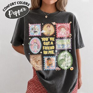 May include: Dark gray Comfort Colors t-shirt featuring a colorful graphic of Toy Story characters in stamp-like designs. The phrase "YOU'VE GOT A FRIEND IN ME" is centered on the shirt. The shirt has the word "Pepper" in a white oval.