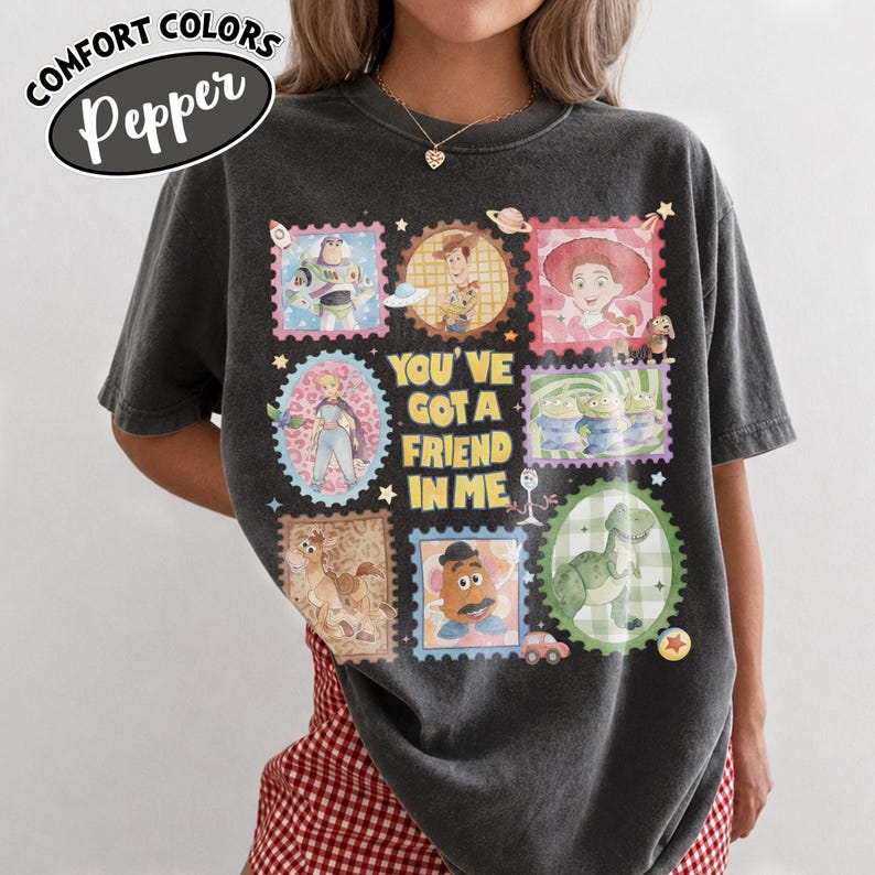 May include: Dark gray Comfort Colors t-shirt featuring a colorful graphic of Toy Story characters in stamp-like designs. The phrase "YOU'VE GOT A FRIEND IN ME" is centered on the shirt. The shirt has the word "Pepper" in a white oval.
