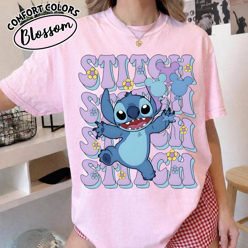 Puede incluir: Camiseta rosa claro con el personaje Stitch de la pel&iacute;cula Lilo & Stitch. La palabra "STITCH" est&aacute; impresa en letras grandes de estilo retro en tonos morados y azules, con detalles de flores amarillas. La camiseta tambi&eacute;n tiene el texto "Comfort Colors Blossom".