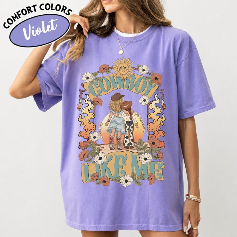 May include: A violet t-shirt featuring a graphic print of two figures in cowboy hats and boots standing together. The design includes a sun, flowers, flame-like shapes, and the text "COWBOY LIKE ME" in a light blue font. A label reads "COMFORT COLORS Violet".