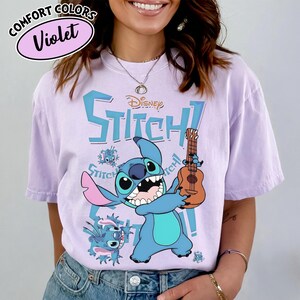 Pu&ograve; includere: Una maglietta Comfort Colors viola con un personaggio dei cartoni animati Stitch che tiene un ukulele. Il design include la scritta "Disney Stitch!" in blu, con grafica Stitch aggiuntiva. La maglietta &egrave; di colore viola chiaro.