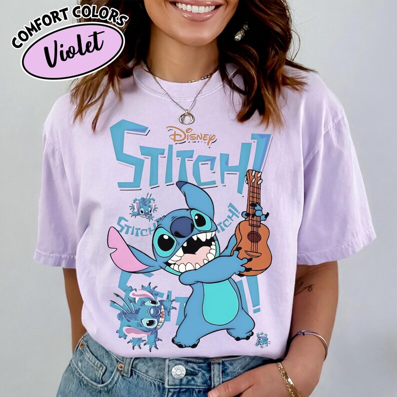 Pu&ograve; includere: Una maglietta Comfort Colors viola con un personaggio dei cartoni animati Stitch che tiene un ukulele. Il design include la scritta "Disney Stitch!" in blu, con grafica Stitch aggiuntiva. La maglietta &egrave; di colore viola chiaro.