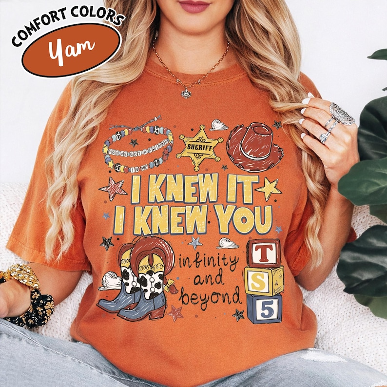 May include: A burnt orange short-sleeve t-shirt featuring a graphic print with a sheriff badge, a cowboy hat, cow-print boots, a beaded bracelet, and three stacked alphabet blocks labelled T, S, and 5. The text reads I KNEW IT I KNEW YOU infinity and beyond.