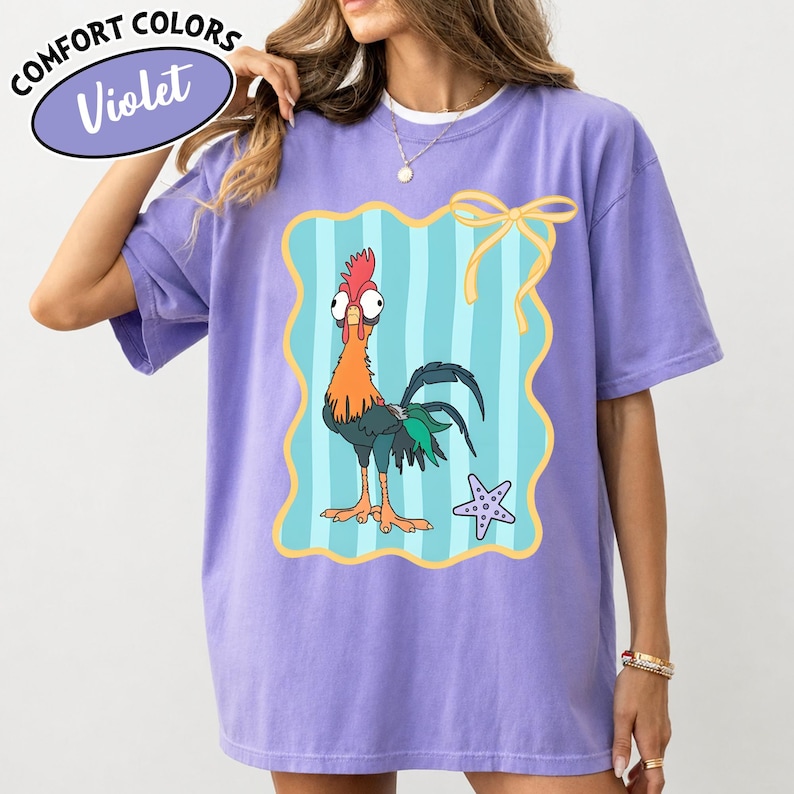 May include: A violet Comfort Colors t-shirt with a cartoon rooster in orange, red, and green. The rooster is set against a light blue striped background with a yellow border and bow. A starfish is also present.