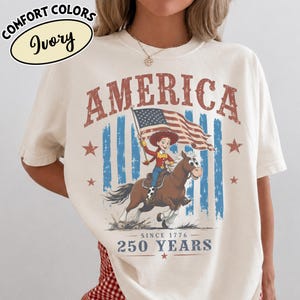 Puede incluir: Una camiseta de color marfil con un gr&aacute;fico de una vaquera montando un caballo marr&oacute;n mientras sostiene una bandera estadounidense. El texto dice AMERICA en la parte superior y SINCE 1776 250 YEARS en la parte inferior. Incluye rayas azules y estrellas rojas.