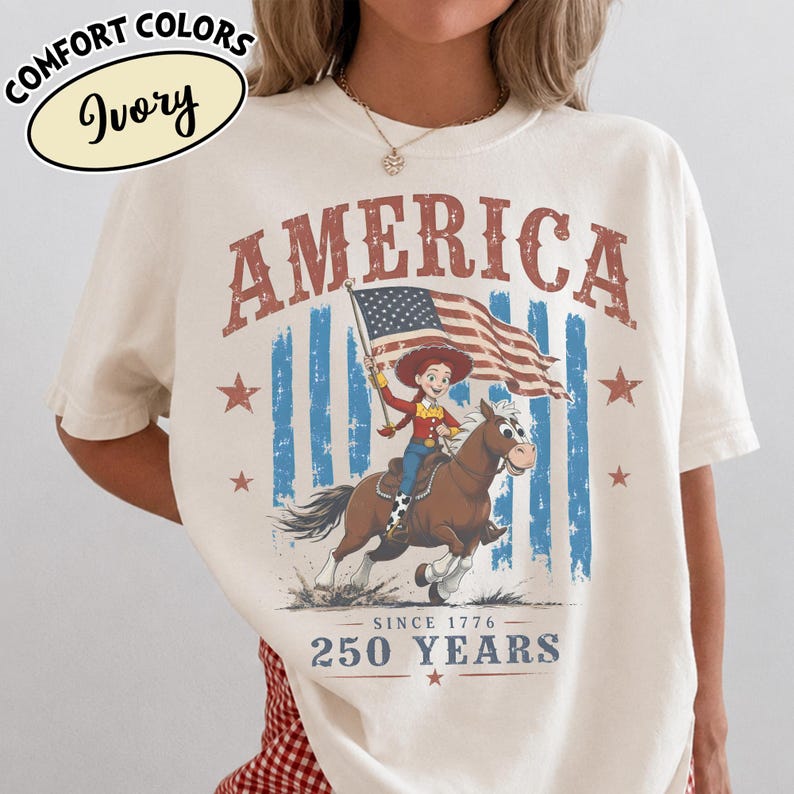 Puede incluir: Una camiseta de color marfil con un gr&aacute;fico de una vaquera montando un caballo marr&oacute;n mientras sostiene una bandera estadounidense. El texto dice AMERICA en la parte superior y SINCE 1776 250 YEARS en la parte inferior. Incluye rayas azules y estrellas rojas.