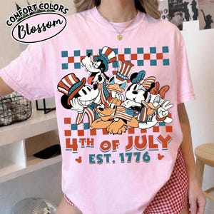 May include: A light pink t-shirt featuring a graphic of cartoon characters wearing patriotic hats and holding American flags. The design includes a red and teal checkered pattern, the text 4TH OF JULY EST. 1776, and small Mickey Mouse head icons.
