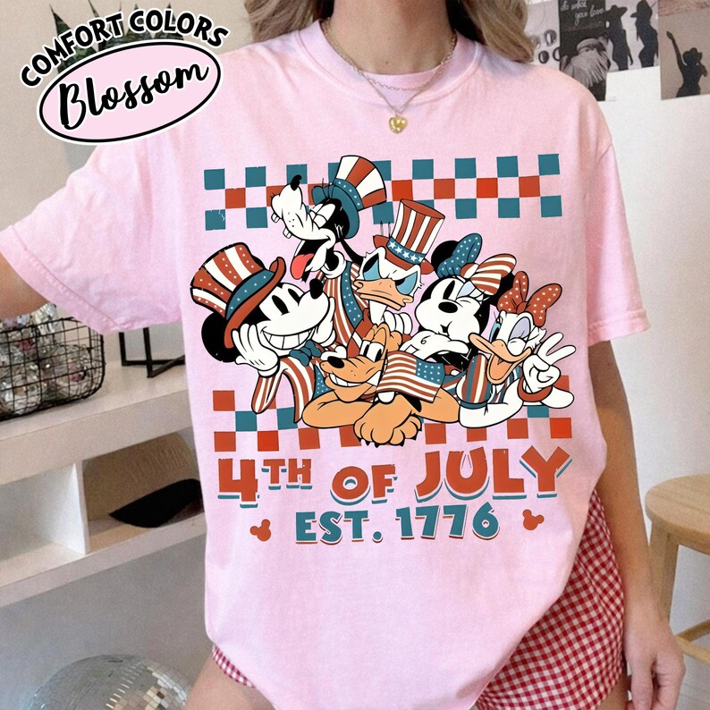 May include: A light pink t-shirt featuring a graphic of cartoon characters wearing patriotic hats and holding American flags. The design includes a red and teal checkered pattern, the text 4TH OF JULY EST. 1776, and small Mickey Mouse head icons.