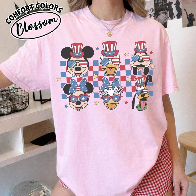 May include: Pink t-shirt with cartoon characters wearing patriotic sunglasses and hats. The shirt features Mickey Mouse, Donald Duck, Pluto, and Minnie Mouse designs. The text "COMFORT COLORS Blossom" is printed on the top left.