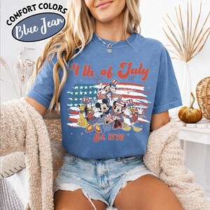 May include: Blue t-shirt with a patriotic design featuring cartoon characters in front of an American flag. The text "4th of July" and "Est. 1776" are in red. The shirt is labeled "Comfort Colors Blue Jean".