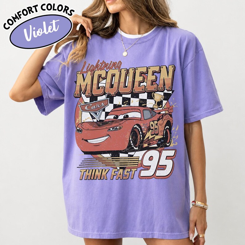 May include: A violet Comfort Colors t-shirt with a graphic of Lightning McQueen from the Cars film. The design includes the text "Lightning McQueen", "Cars", "95", and "Think Fast". The shirt has a crew neck and short sleeves.