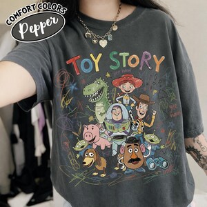 May include: Dark grey t-shirt featuring a colorful graphic of Toy Story characters, including Woody, Buzz Lightyear, and Rex. The words "TOY STORY" are written in rainbow colors. The shirt has a relaxed fit and the words "Comfort Colors Pepper" are in the upper left corner.