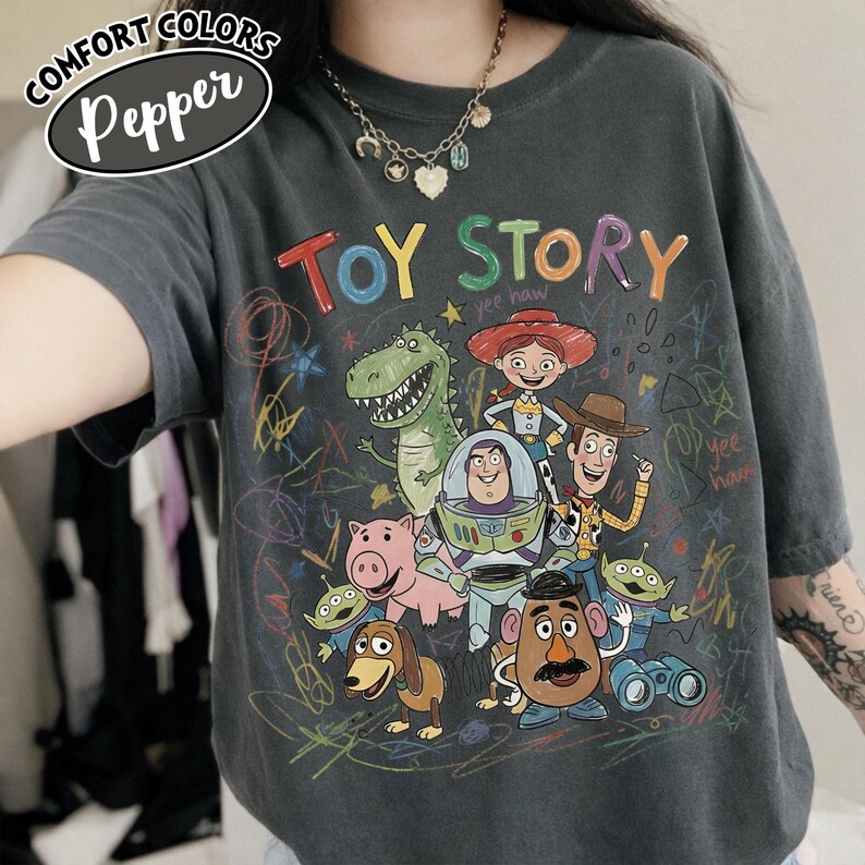 May include: Dark grey t-shirt featuring a colorful graphic of Toy Story characters, including Woody, Buzz Lightyear, and Rex. The words "TOY STORY" are written in rainbow colors. The shirt has a relaxed fit and the words "Comfort Colors Pepper" are in the upper left corner.