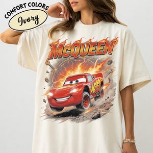 May include: Ivory-colored t-shirt featuring a graphic of Lightning McQueen from the movie Cars. The design includes the word "McQueen" in fiery letters, the number 95, and a racing scene with sparks and tools.
