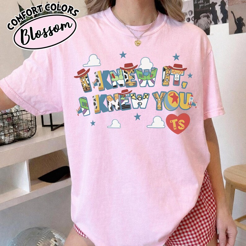 May include: A light pink short-sleeve t-shirt featuring the text I KNEW IT, I KNEW YOU in a colorful, patterned font. The design includes small cowboy hats, stars, clouds, and a red heart with the letters TS. The shirt has a crew neckline.