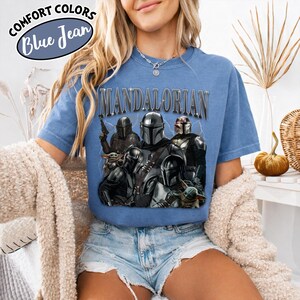 Puede incluir: Camiseta azul de manga corta con la palabra "MANDALORIAN" en letras plateadas. La camiseta presenta un gr&aacute;fico de varios personajes de la serie Star Wars, incluyendo a The Mandalorian y Grogu. La camiseta tiene las palabras "COMFORT COLORS" y "Blue Jean" en un c&iacute;rculo.