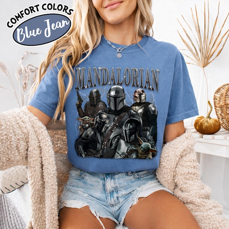 Puede incluir: Camiseta azul de manga corta con la palabra "MANDALORIAN" en letras plateadas. La camiseta presenta un gr&aacute;fico de varios personajes de la serie Star Wars, incluyendo a The Mandalorian y Grogu. La camiseta tiene las palabras "COMFORT COLORS" y "Blue Jean" en un c&iacute;rculo.