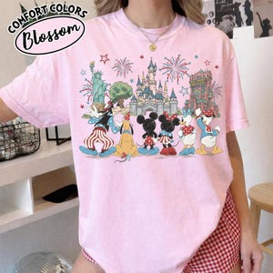 以下が含まれることがあります： ディズニーのキャラクターとランドマーク、自由の女神や城などのカラフルなグラフィックが特徴のライトピンクのTシャツ。「Comfort Colors Blossom」の文字が円形にデザインされています。