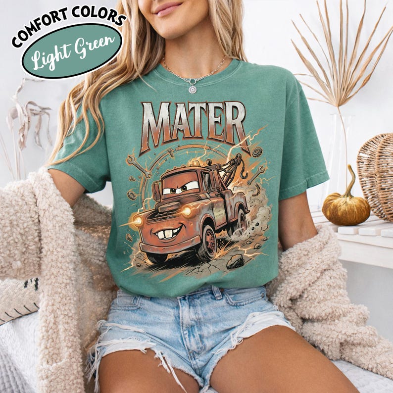 Puede incluir: Camiseta verde claro con una imagen de dibujos animados de Mate de la pel&iacute;cula Cars. El gr&aacute;fico incluye la palabra "MATER" en naranja. La camiseta tambi&eacute;n tiene las palabras "COMFORT COLORS" y "Light Green" en un dise&ntilde;o circular.