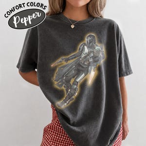 Puede incluir: Una camiseta gris carb&oacute;n de manga corta con un estampado gr&aacute;fico de un personaje blindado que sostiene un bl&aacute;ster y lleva una peque&ntilde;a criatura. El texto COMFORT COLORS Pepper est&aacute; impreso en la esquina superior izquierda. Cuello redondo.