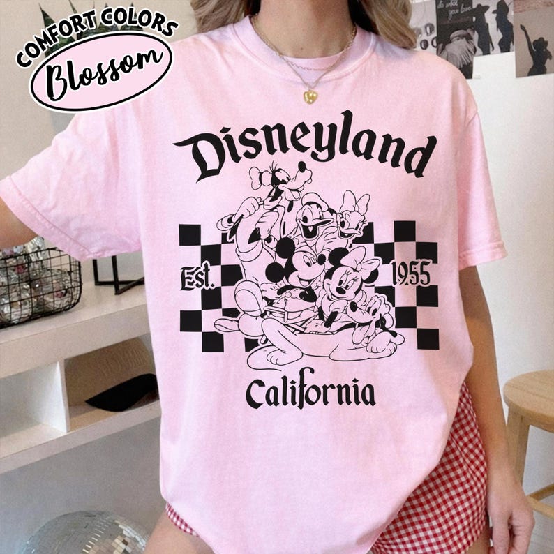 May include: A light pink t-shirt featuring the word "Disneyland" in a stylized font, along with a graphic of Mickey Mouse, Minnie Mouse, Goofy, Donald Duck, and Pluto. The shirt also includes the text "California" and "Est. 1955".