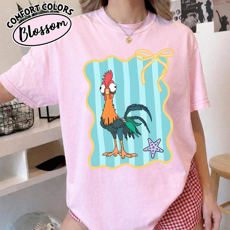 May include: A pale pink t-shirt with a cartoon rooster design inside a blue and yellow frame. The rooster is orange, red, and green. The shirt also has the words "Comfort Colors Blossom" in a circular design.