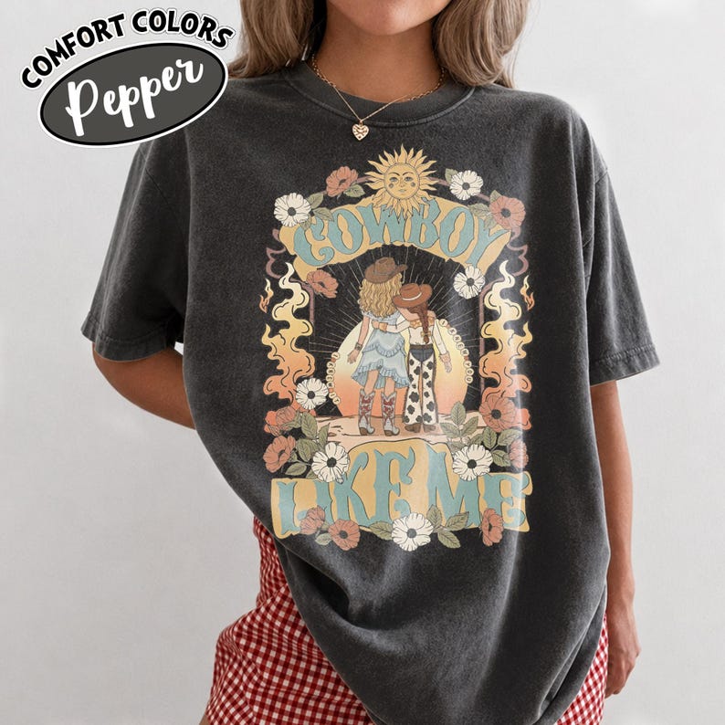May include: A charcoal gray t-shirt featuring a graphic print of two figures in cowboy hats and boots standing before a sun. The text reads COWBOY LIKE ME. The design is surrounded by flowers and flames in shades of orange, blue, and white.