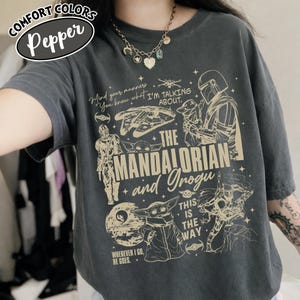 Op de afbeelding: Donkergrijs Comfort Colors T-shirt met een afbeelding van The Mandalorian en Grogu. Het ontwerp bevat illustraties van personages, schepen en de tekst "Mind your manners, You know what I'm talking about." en "This is the way."