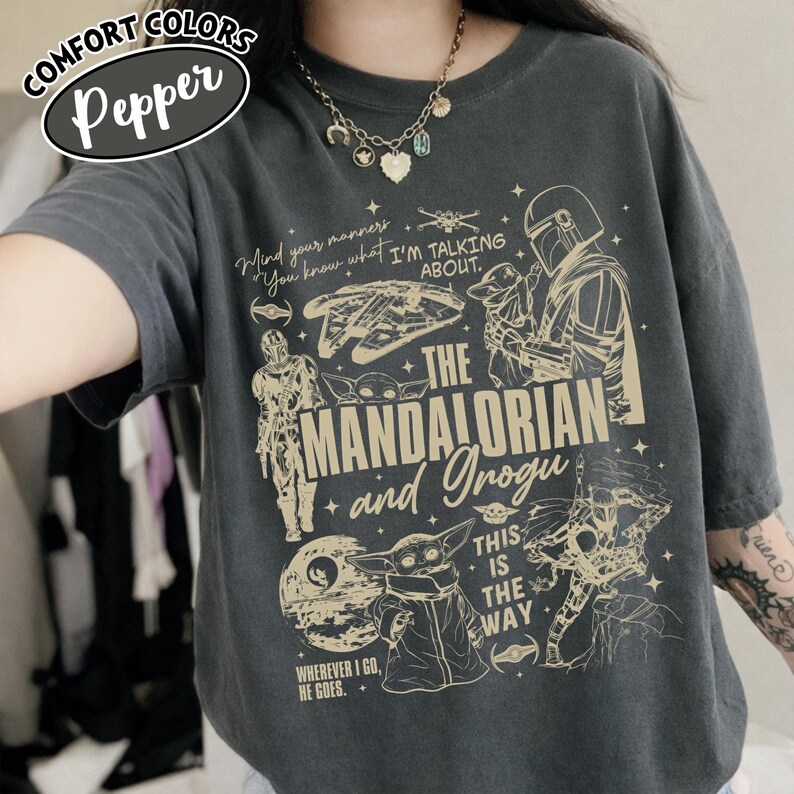 Op de afbeelding: Donkergrijs Comfort Colors T-shirt met een afbeelding van The Mandalorian en Grogu. Het ontwerp bevat illustraties van personages, schepen en de tekst "Mind your manners, You know what I'm talking about." en "This is the way."