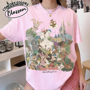 Op de afbeelding: Roze T-shirt met een afbeelding van verschillende Disney-personages, waaronder Mickey Mouse, Dumbo en Peter Pan. Op het shirt staan de woorden "Magic Kingdom" en "Walt Disney World". In de linkerbovenhoek staan de woorden "Comfort Colors" en "Blossom".