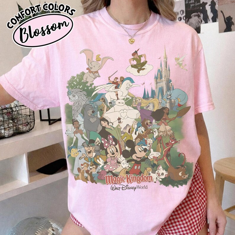 Op de afbeelding: Roze T-shirt met een afbeelding van verschillende Disney-personages, waaronder Mickey Mouse, Dumbo en Peter Pan. Op het shirt staan de woorden "Magic Kingdom" en "Walt Disney World". In de linkerbovenhoek staan de woorden "Comfort Colors" en "Blossom".