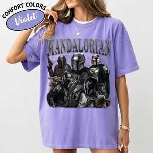 Puede incluir: Camiseta violeta Comfort Colors con un gr&aacute;fico de personajes de Mandalorian. La palabra "MANDALORIAN" est&aacute; impresa en la parte superior. La camiseta tiene cuello redondo y mangas cortas.