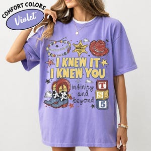 May include: A violet short-sleeve t-shirt featuring a graphic print with a sheriff badge, a cowboy hat, cow-print boots, a beaded bracelet, and alphabet blocks. The text reads I KNEW IT I KNEW YOU infinity and beyond. The shirt has a crew neckline.