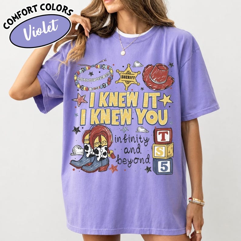 May include: A violet short-sleeve t-shirt featuring a graphic print with a sheriff badge, a cowboy hat, cow-print boots, a beaded bracelet, and alphabet blocks. The text reads I KNEW IT I KNEW YOU infinity and beyond. The shirt has a crew neckline.
