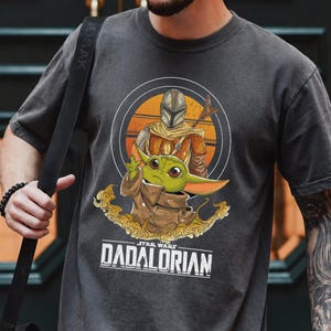 Może przedstawiać: Ciemnoszara koszulka z grafiką Mandalorianina i Grogu z Gwiezdnych Wojen. Grogu pokazuje znak pokoju. Tekst "DADALORIAN" jest wydrukowany pod obrazem. Tło ma pomarańczowo-ż&oacute;łty wz&oacute;r zachodu słońca.