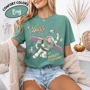 Może przedstawiać: Zielona koszulka z Buzzem Astralem, napisem "Buzz Lightyear" i "To Infinity and Beyond". Koszulka ma stylizowany design z planetą i rakietą.