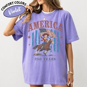 Puede incluir: Una camiseta violeta de manga corta con un estampado gr&aacute;fico que incluye el texto AMERICA, SINCE 1776 y 250 YEARS. El dise&ntilde;o muestra un personaje de dibujos animados montando un caballo marr&oacute;n y sosteniendo una bandera estadounidense, rodeado de estrellas y rayas azules.