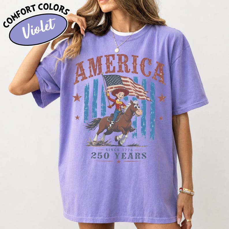 Puede incluir: Una camiseta violeta de manga corta con un estampado gr&aacute;fico que incluye el texto AMERICA, SINCE 1776 y 250 YEARS. El dise&ntilde;o muestra un personaje de dibujos animados montando un caballo marr&oacute;n y sosteniendo una bandera estadounidense, rodeado de estrellas y rayas azules.