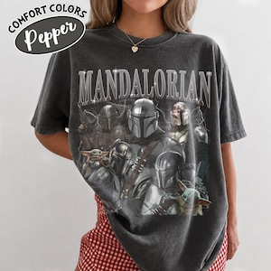 Puede incluir: Camiseta gris oscuro con la palabra "MANDALORIAN" en fuente plateada desgastada. La camiseta presenta un gr&aacute;fico de m&uacute;ltiples personajes Mandalorianos y Grogu. La camiseta tiene las palabras "Comfort Colors" y "Pepper" en un &oacute;valo blanco.