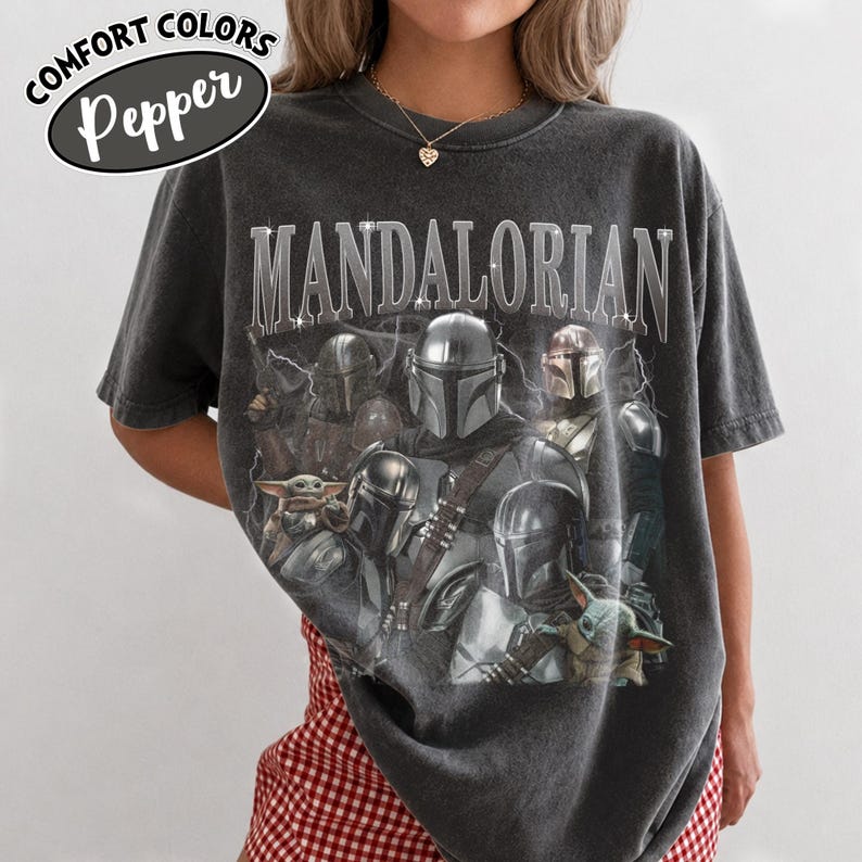 Puede incluir: Camiseta gris oscuro con la palabra "MANDALORIAN" en fuente plateada desgastada. La camiseta presenta un gr&aacute;fico de m&uacute;ltiples personajes Mandalorianos y Grogu. La camiseta tiene las palabras "Comfort Colors" y "Pepper" en un &oacute;valo blanco.