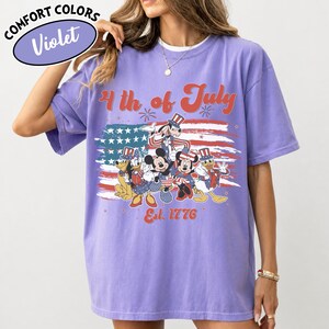 May include: Violet Comfort Colors t-shirt featuring a patriotic design with Mickey Mouse, Minnie Mouse, Donald Duck, Goofy, and Pluto in front of an American flag. The text reads "4th of July" and "Est. 1776".