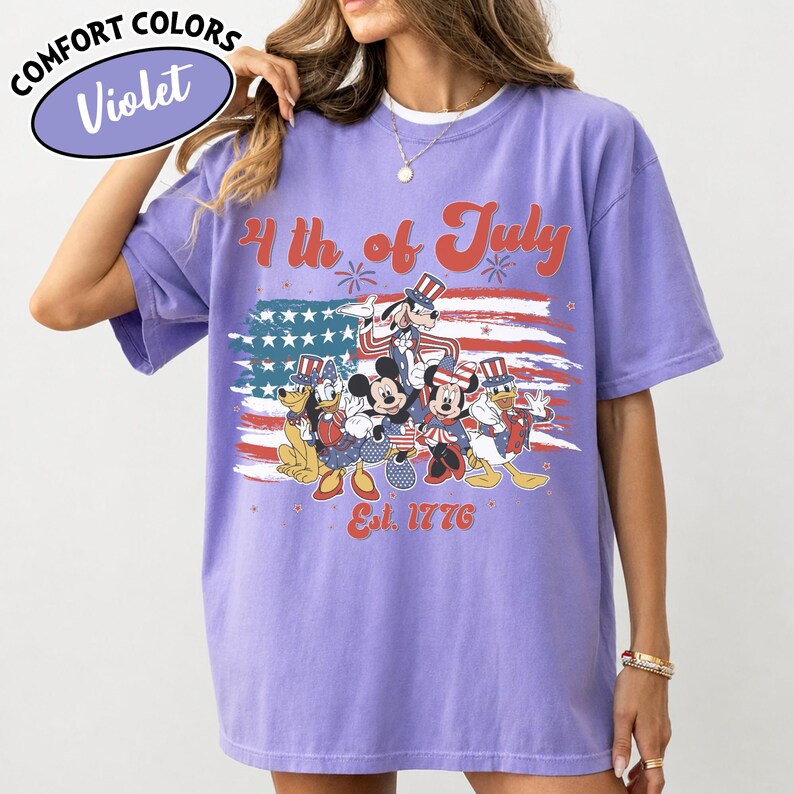 May include: Violet Comfort Colors t-shirt featuring a patriotic design with Mickey Mouse, Minnie Mouse, Donald Duck, Goofy, and Pluto in front of an American flag. The text reads "4th of July" and "Est. 1776".