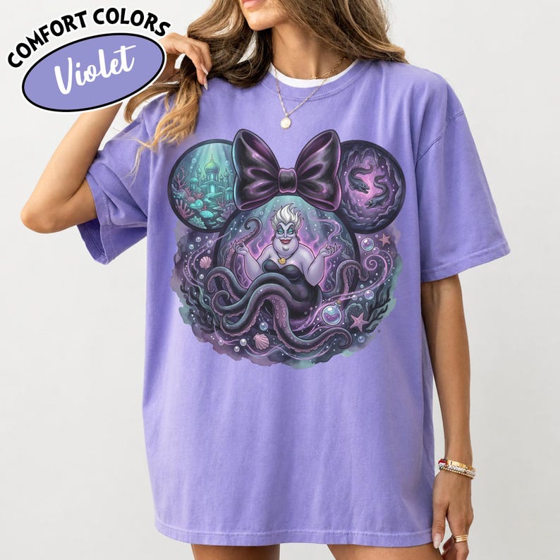 May include: A violet-colored t-shirt featuring a graphic of Ursula from The Little Mermaid, set within a Mickey Mouse ear design. The design includes a large bow, underwater scenes, and the text "Comfort Colors Violet".