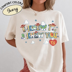 May include: An ivory t-shirt featuring the text I KNEW IT, I KNEW YOU in a multi-colored graphic font inspired by toy characters. The design includes small blue stars, white cloud shapes, and a red heart with the letters TS inside the heart shape.