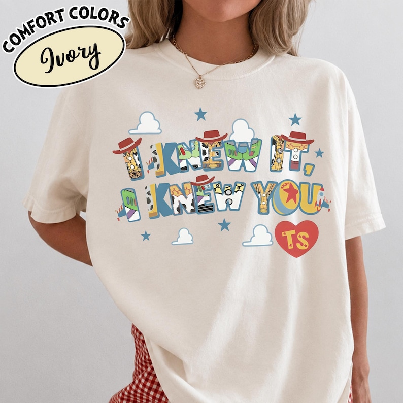 May include: An ivory t-shirt featuring the text I KNEW IT, I KNEW YOU in a multi-colored graphic font inspired by toy characters. The design includes small blue stars, white cloud shapes, and a red heart with the letters TS inside the heart shape.