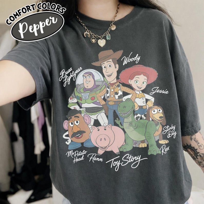 May include: Dark gray Comfort Colors t-shirt featuring a graphic of Toy Story characters including Woody, Buzz Lightyear, and Jessie. The shirt has the text "Toy Story" and "Comfort Colors Pepper" printed on it.