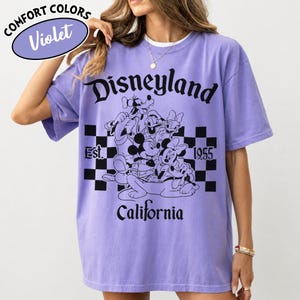 May include: Violet Comfort Colors t-shirt featuring a black and white graphic of Disney characters, including Mickey and Minnie Mouse, Goofy, and Donald Duck. The shirt has the Disneyland logo, "Est. 1955," and "California" printed on it.